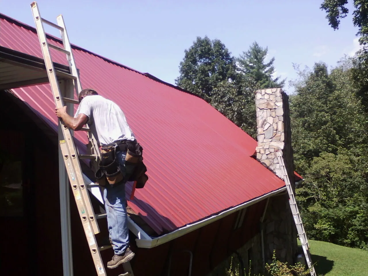 Roofer on a ladder beside a red metal roof and stone chimney — Soffit & Fascia Repair by Roofing Brothers in New Milford